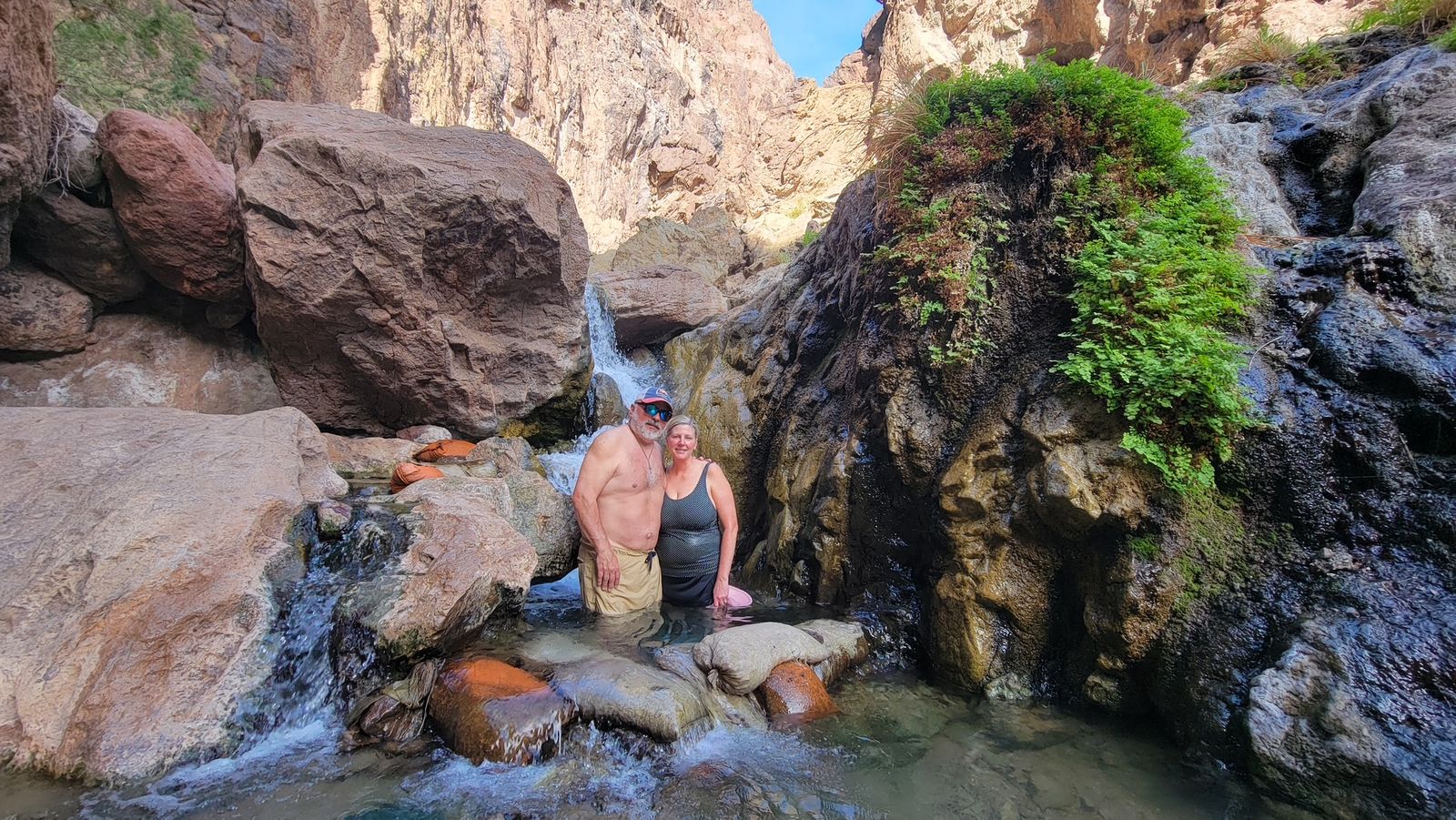 Two people standing in a rocky stream with a small waterfall and lush green plants.
