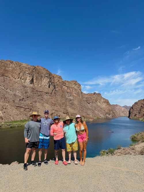 Five people in summer clothes pose by a river with rocky cliffs under a clear blue sky.