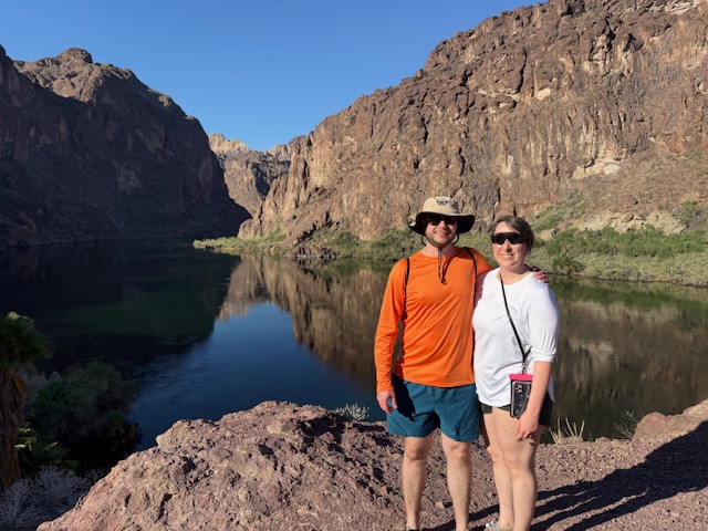 Two people posing by a lake with rocky cliffs under a clear blue sky.