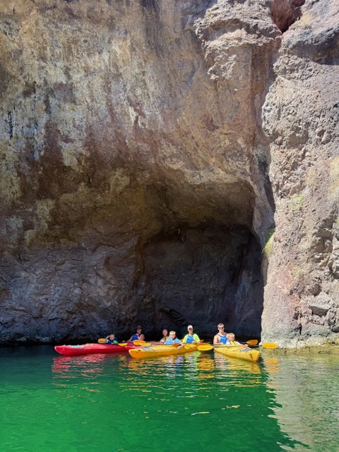 People kayaking under a rocky cliff near a calm green lake.