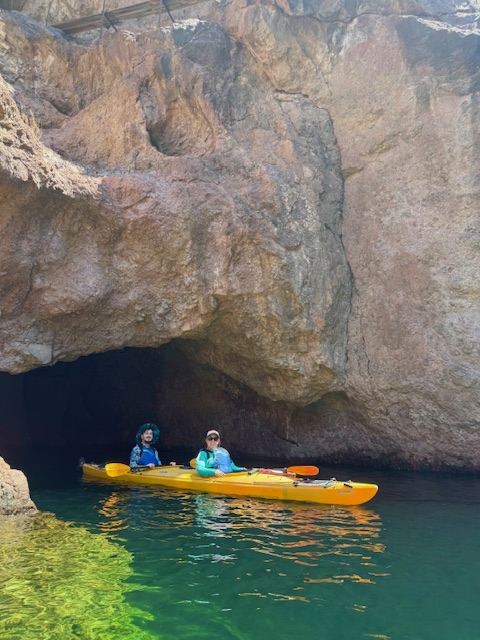 Two people kayaking near a rocky cave entrance over green water.