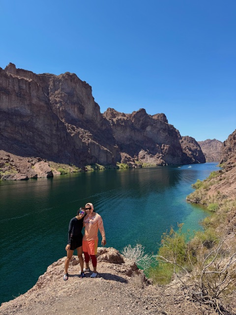 Two people stand on a rocky cliff, overlooking a river with cliffs under a clear blue sky.
