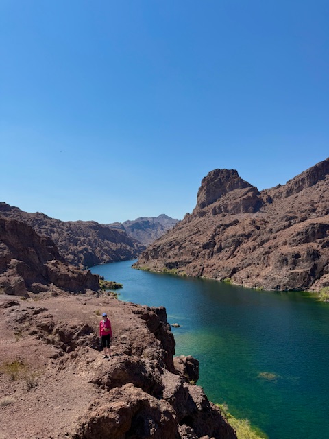 Person in pink hiking along a rocky canyon with a river under a clear blue sky.