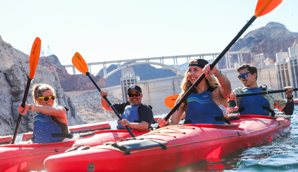 Four people kayaking with paddles, wearing life jackets and sunglasses, near a dam on a sunny day.