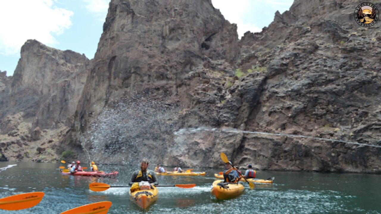 Group kayaking on a river with rocky cliffs in the background, water splashing nearby.
