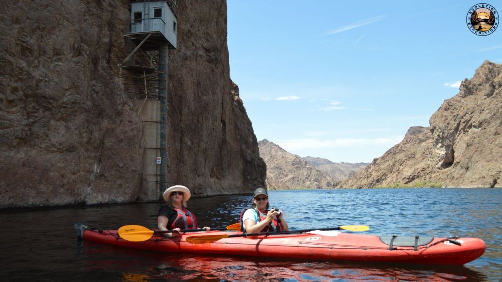 Two people in a red kayak on a river near rocky cliffs under a clear blue sky.