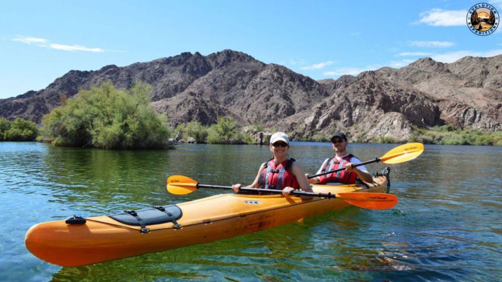 Two people in a kayak on a calm lake with rocky mountains in the background.