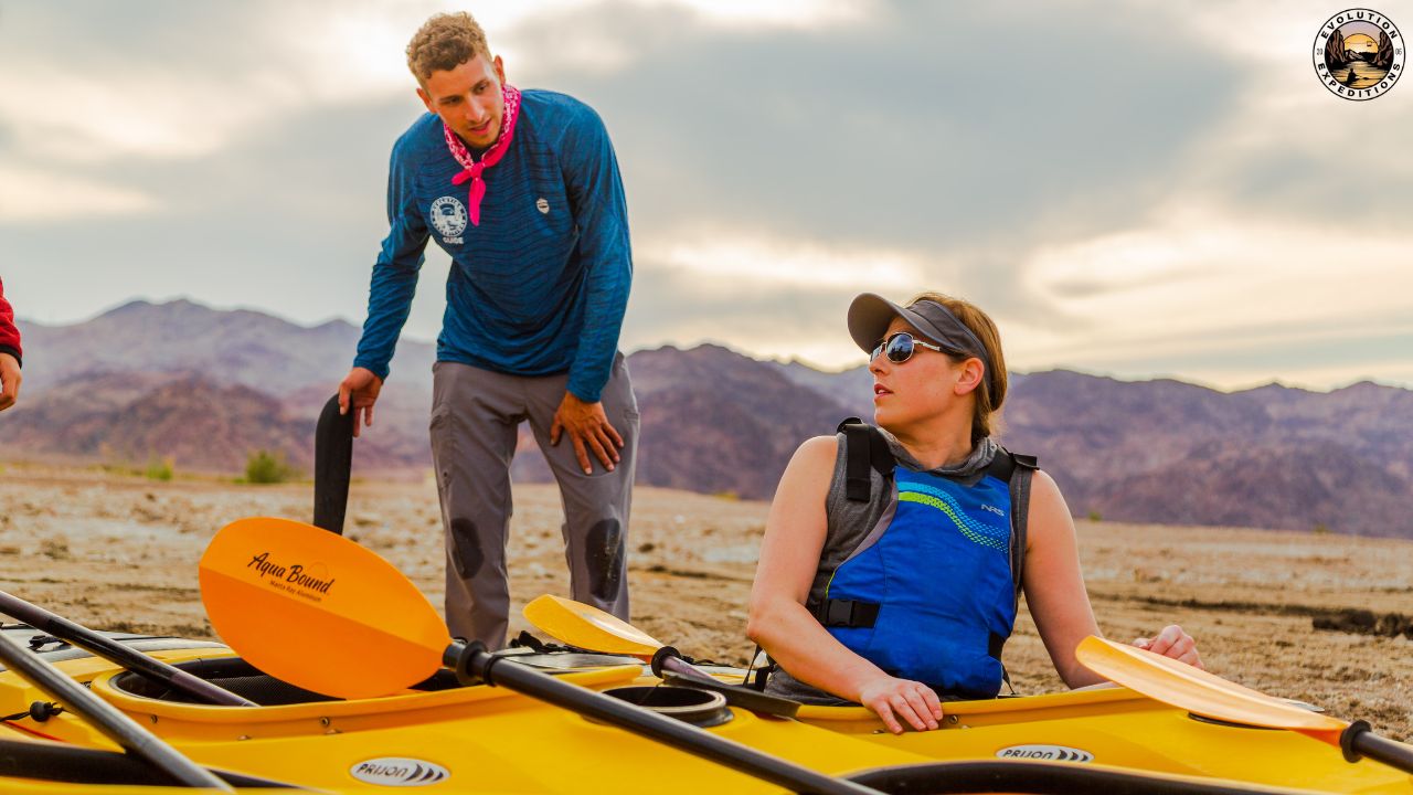 Two people preparing kayaks on a sandy beach with mountains in the background.