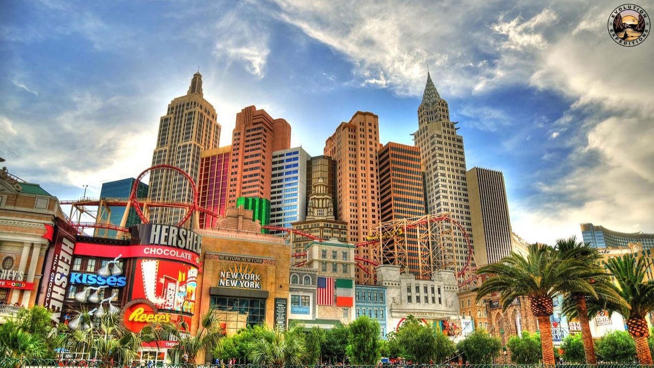 Colorful hotel facade with skyscraper replicas, palm trees, and a roller coaster under a bright sky.