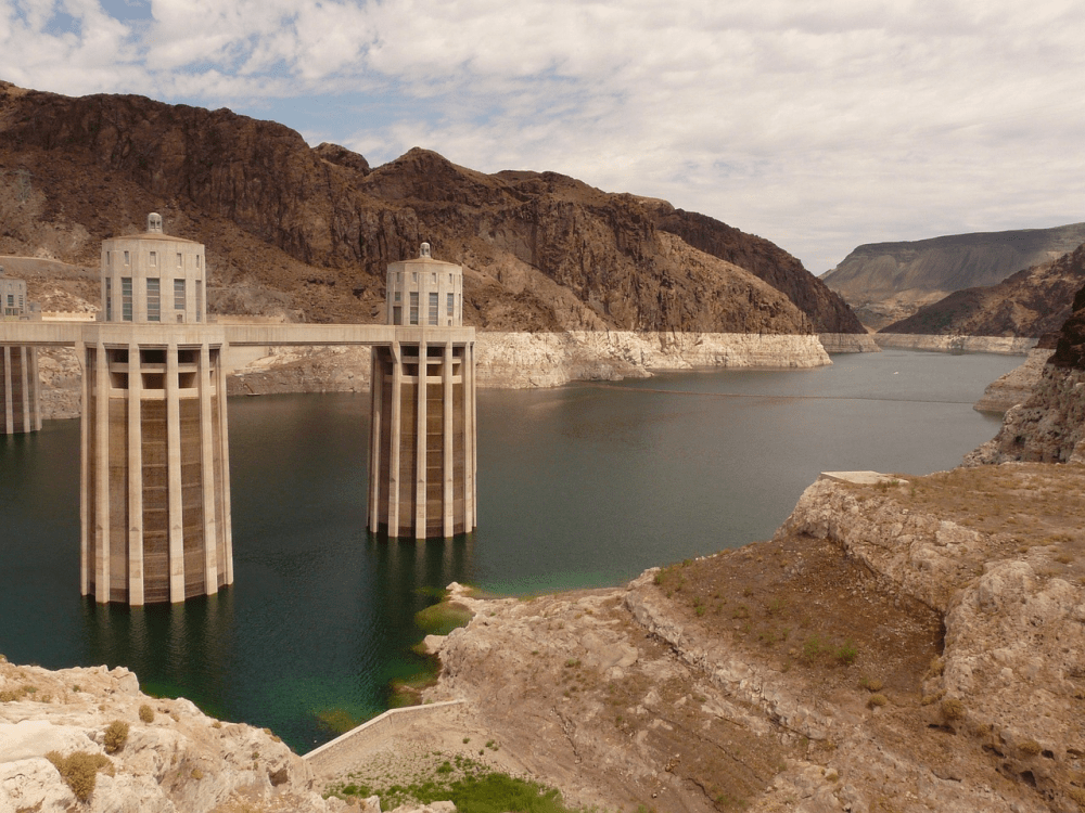 Concrete dam with two towers in front of rocky hills and a body of water.