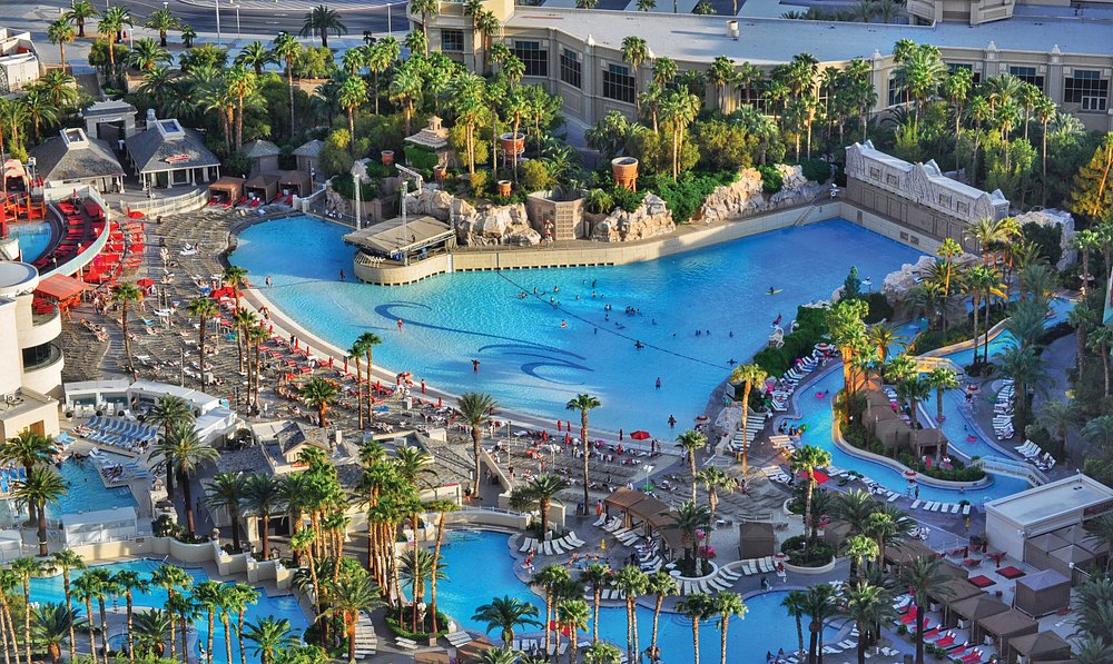 Aerial view of a large, vibrant hotel pool area with palm trees and lounge chairs.