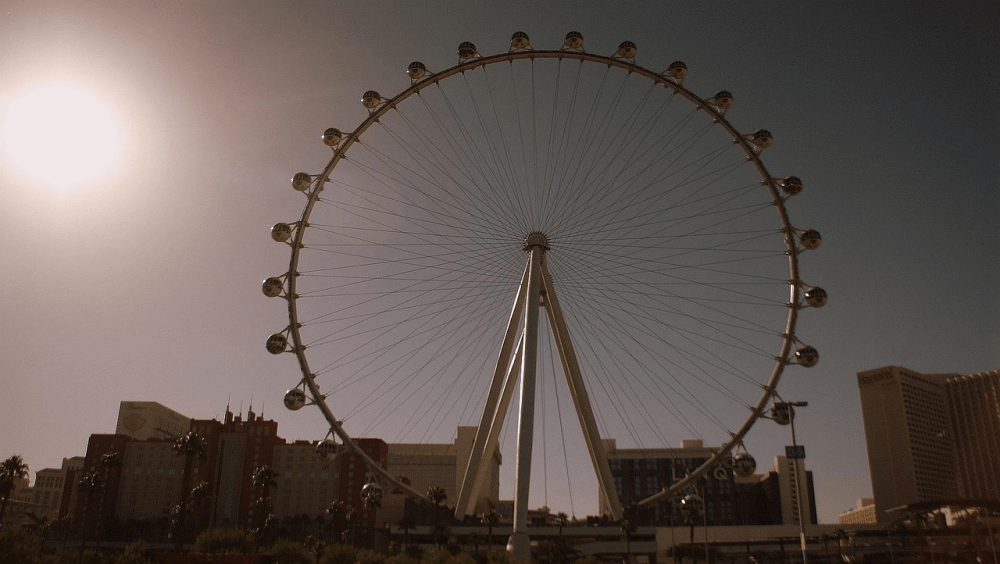 Large Ferris wheel with city buildings and bright sun in the background.