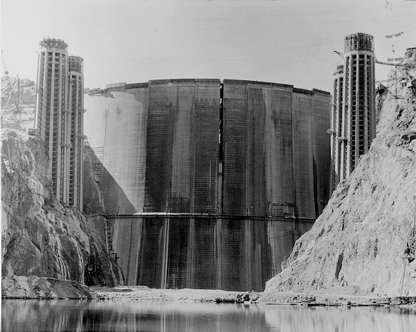 Black and white image of a large dam under construction between two cliffs over a river.