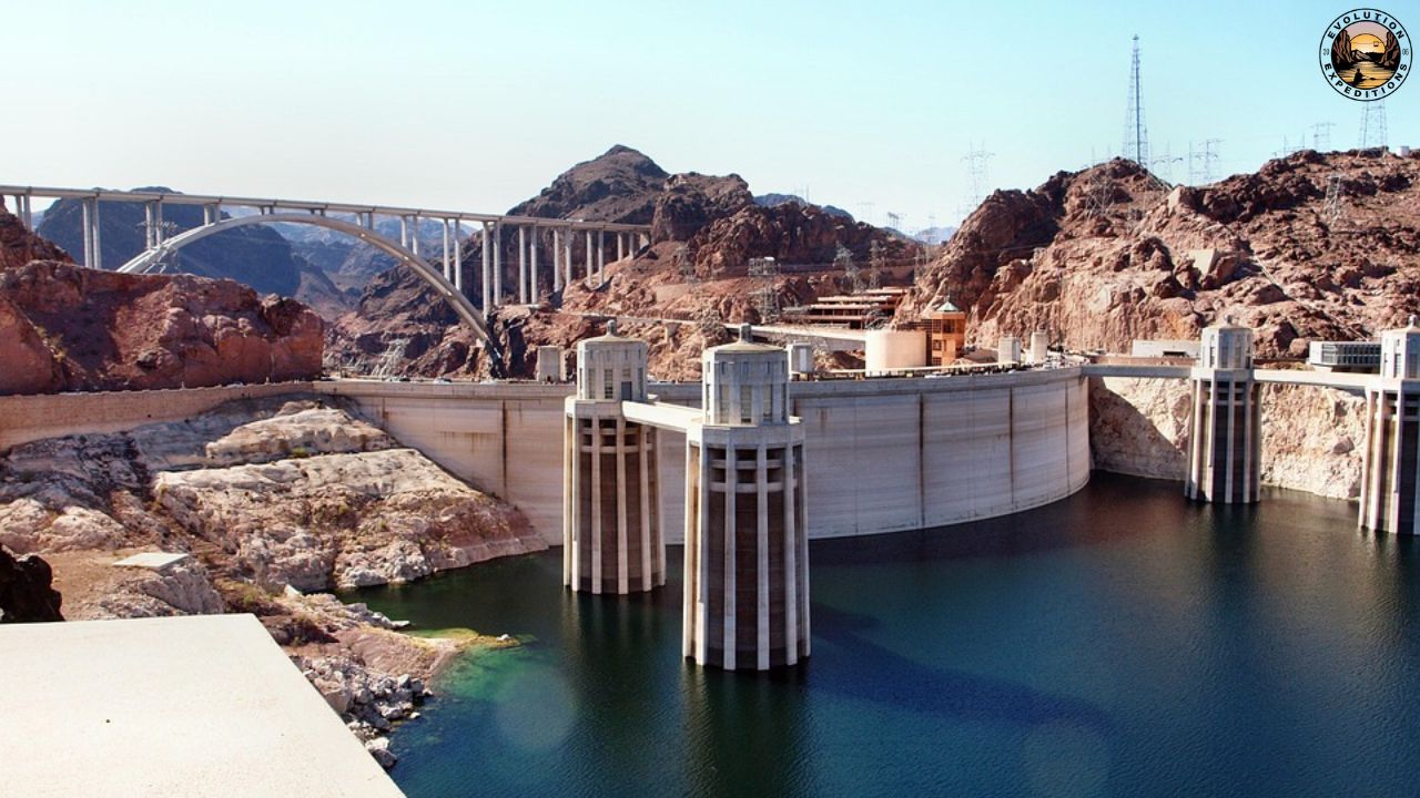 Large dam with water reservoir, arched bridge, and rocky landscape in background.