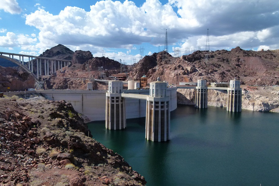 Wide view of a large dam with towers and surrounding rocky landscape under a partly cloudy sky.