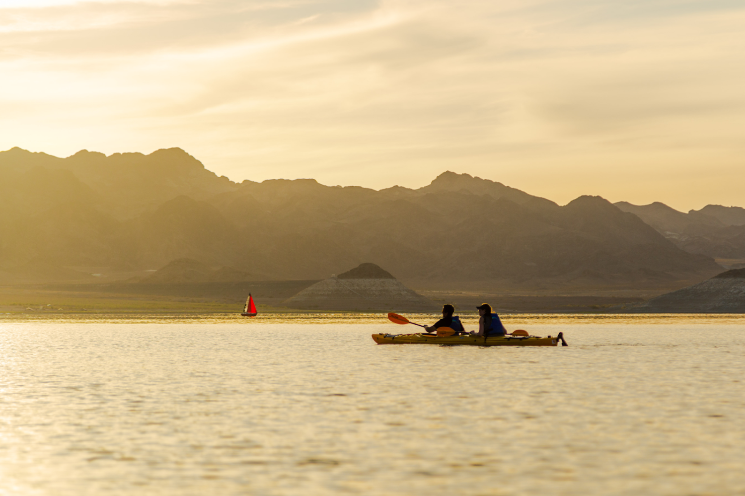 Two people kayaking on a lake at sunset with mountains in the background and a sailboat nearby.