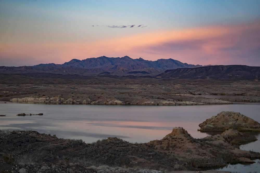 Desert landscape with a calm lake, distant mountains, and a vibrant sunset sky.