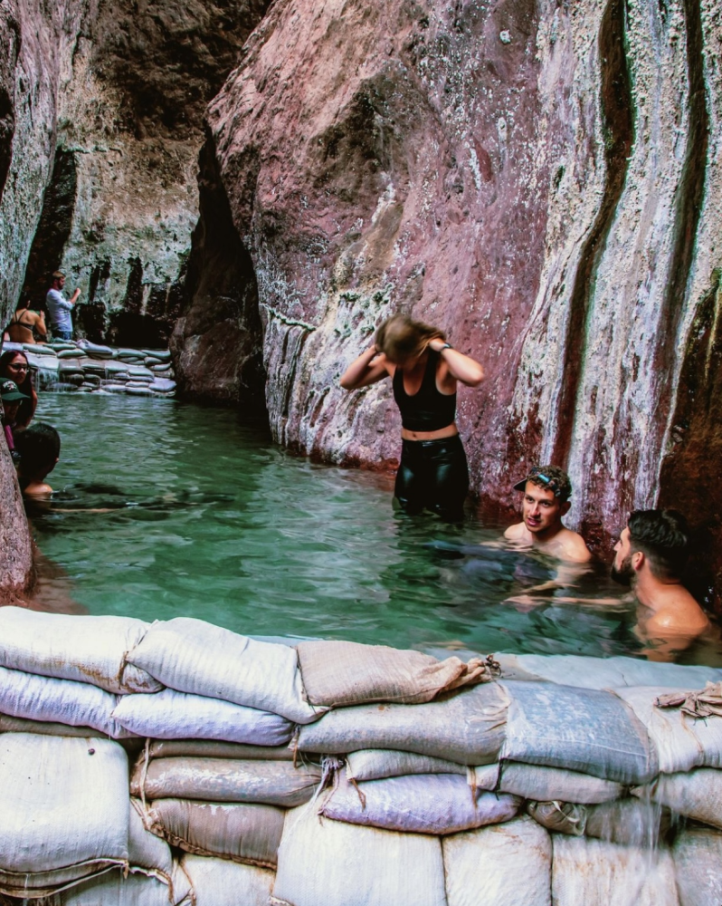 People in a rocky canyon with water, sandbags in foreground; one person adjusts hair, others swim.