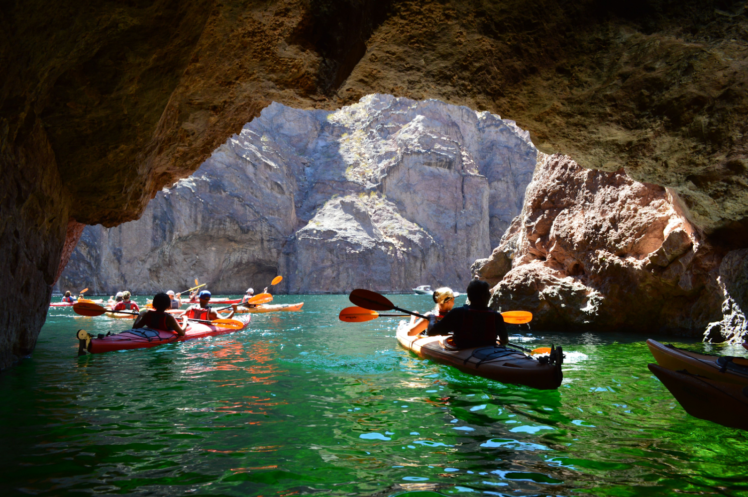 Kayakers paddling through a cave by a rocky waterway during daylight.
