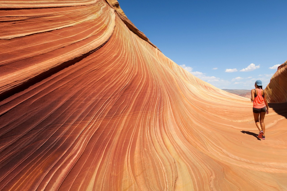 Person in orange tank top walking on wavy sandstone formation under blue sky.