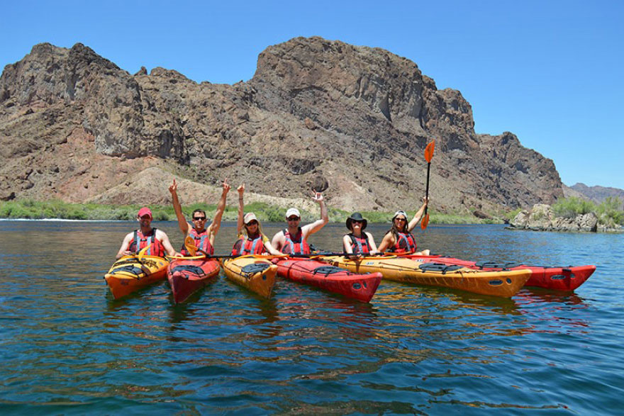 Five people in kayaks on a lake with rocky hills in the background, raising paddles in celebration.