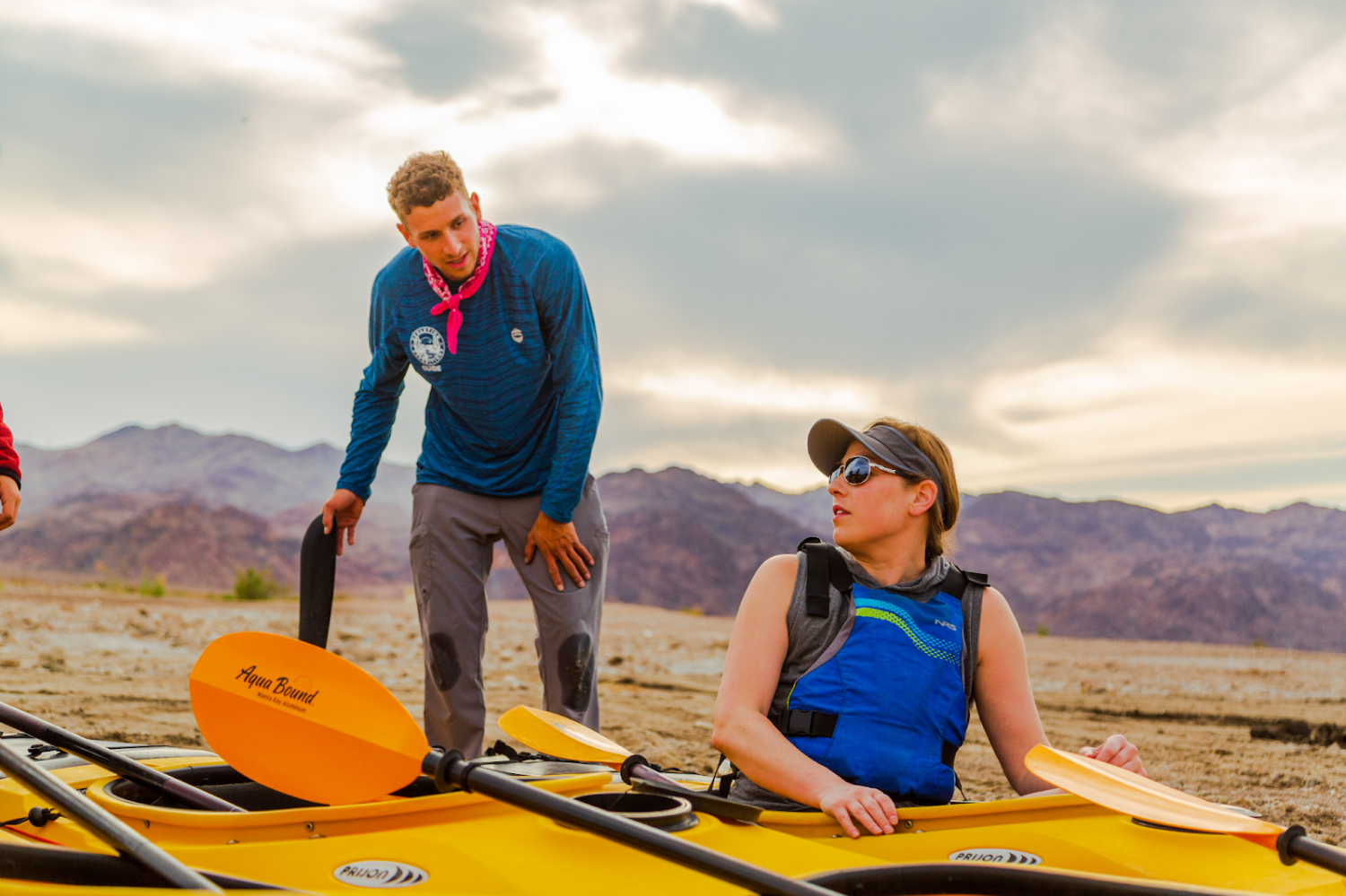 Two people with kayaks on a sandy beach with mountains in the background.