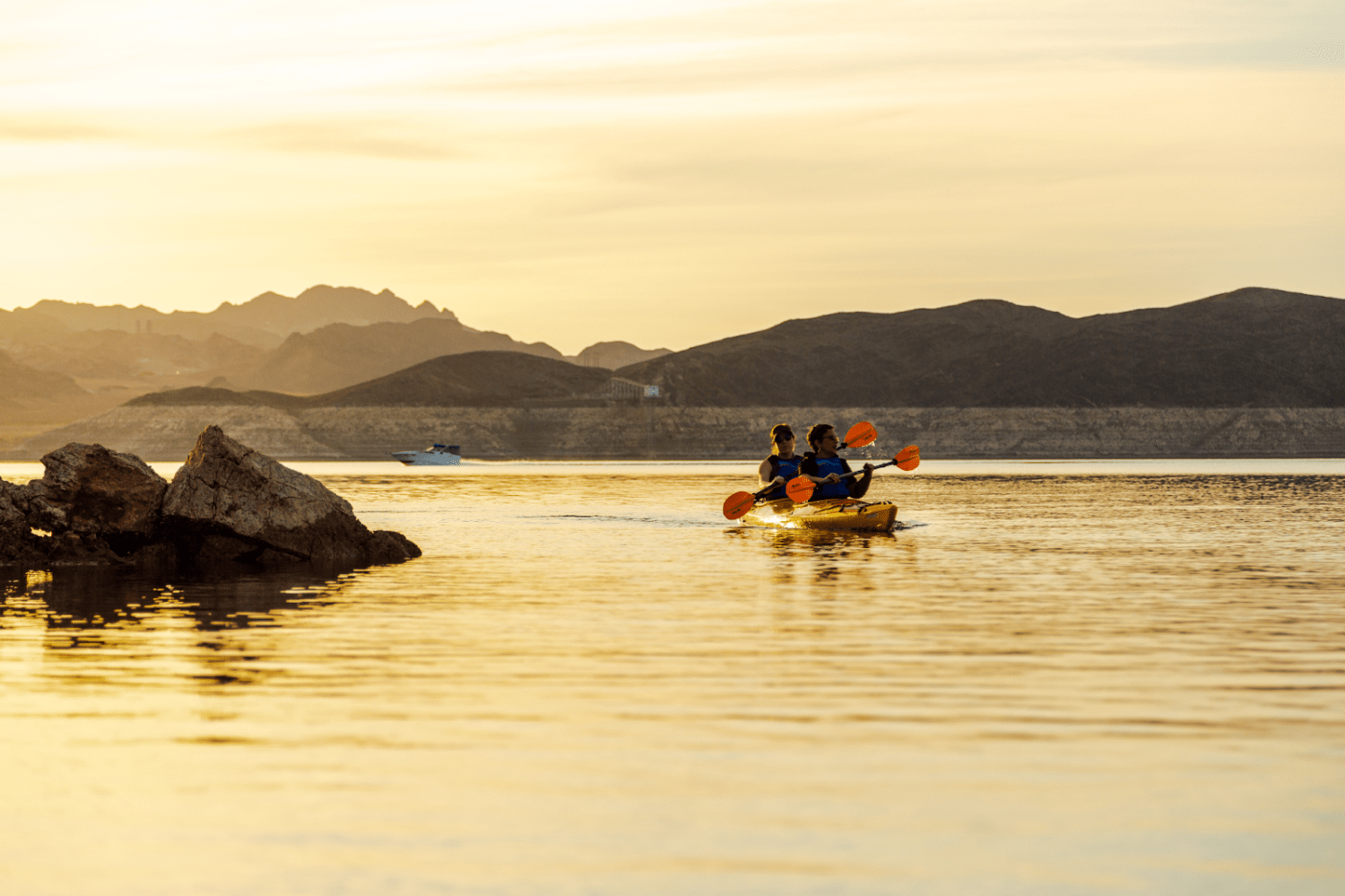 Three people kayaking on a calm lake at sunset with mountains in the background.