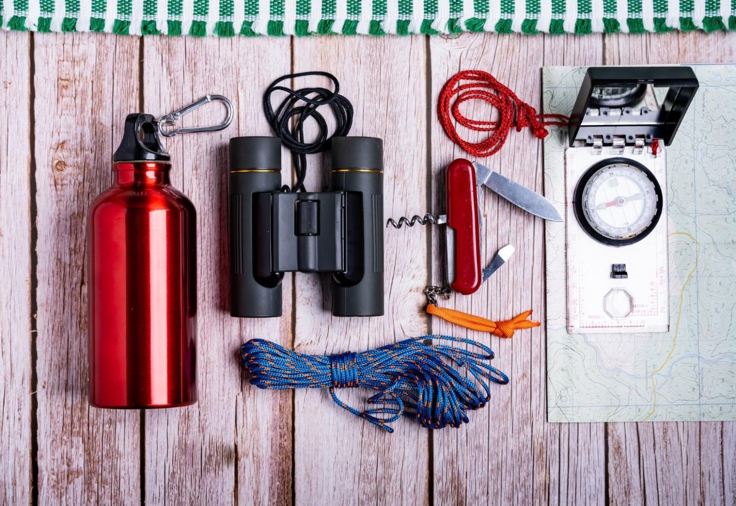 Camping gear on wooden surface: water bottle, binoculars, rope, knife, compass, and map.