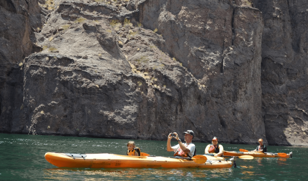 People kayaking in orange kayaks on a green river by rocky cliffs.