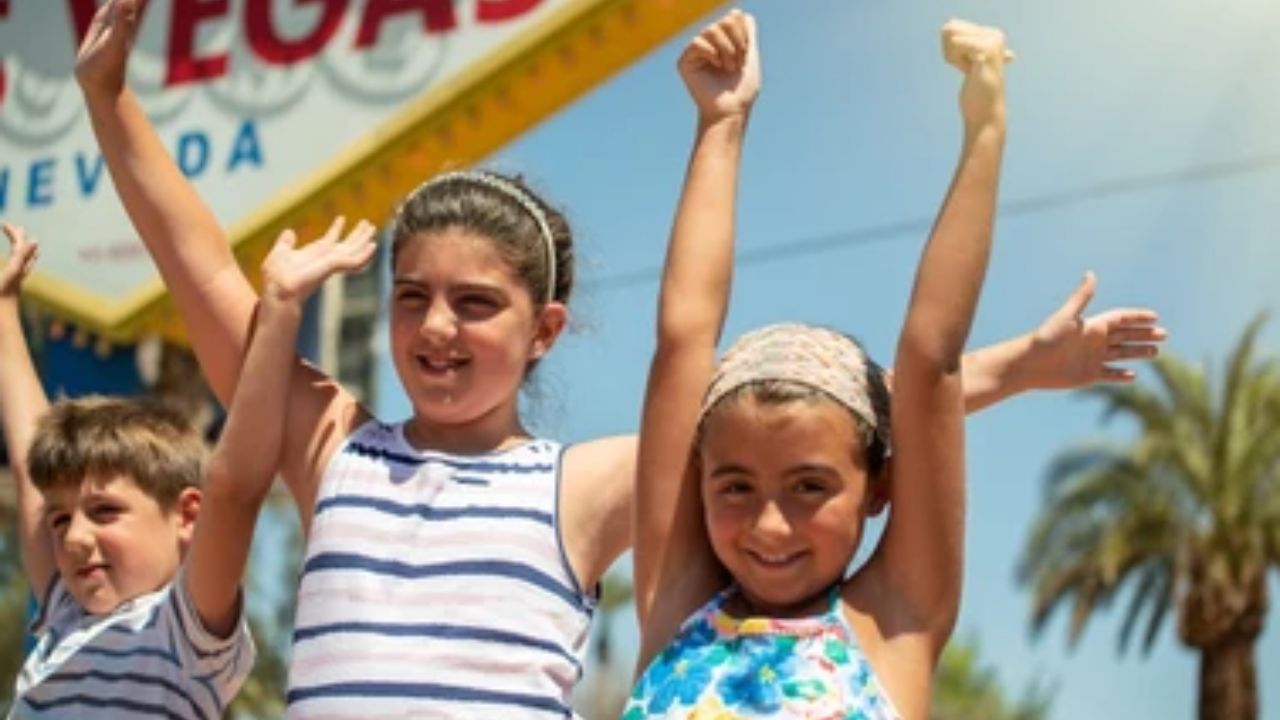 Three children raising arms in front of the Las Vegas sign on a sunny day.