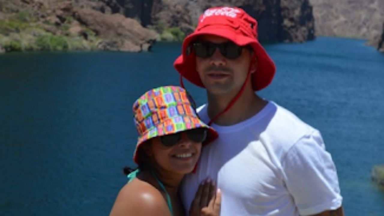 Couple in colorful hats and sunglasses by a lake with rocky cliffs.