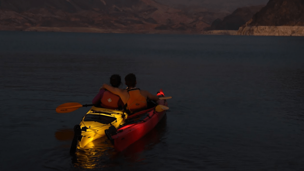 Two people in kayaks on calm water at dusk with distant mountains.