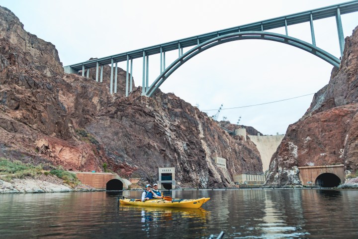 Kayakers on a river under a large bridge, with rocky cliffs and a dam in the background.