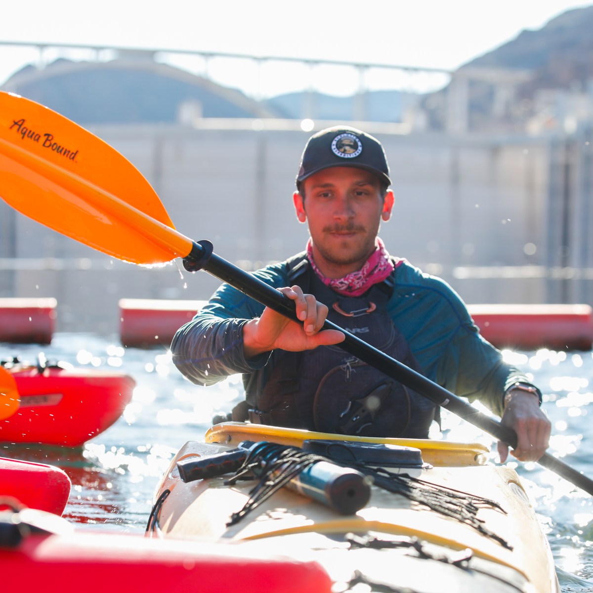 Man kayaking on a sunny day with an orange paddle, wearing a cap and neck scarf.