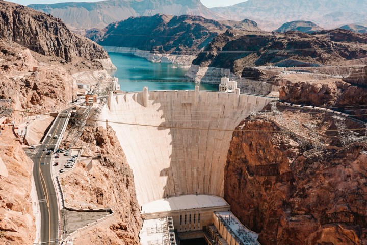 A large dam between rocky cliffs with a lake and clear blue sky in the background.