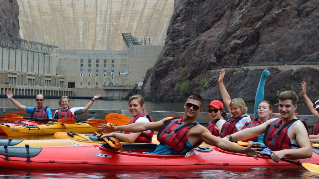 Group of people in kayaks wearing life vests, posing with a large dam in the background.