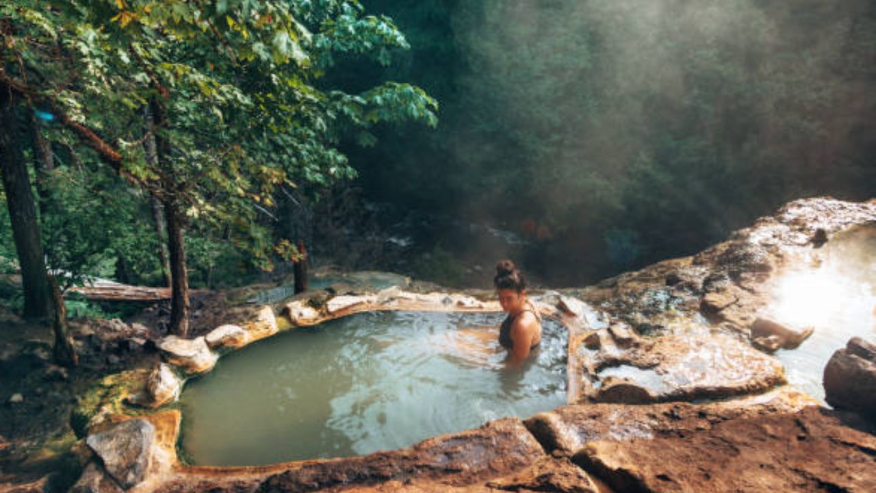 Person soaking in a natural outdoor hot spring pool surrounded by rock formations and forest trees.