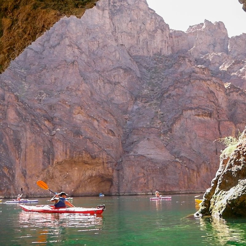 Kayakers paddle in a rocky canyon's calm river beneath towering cliffs.