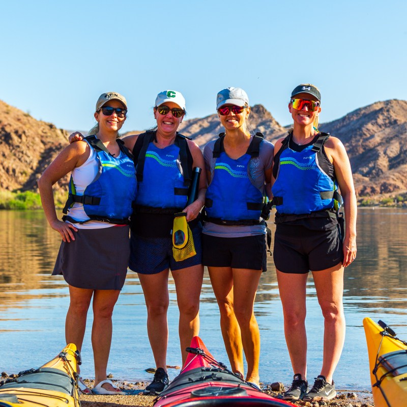 Four people in blue life vests stand by kayaks near a lake with mountains.