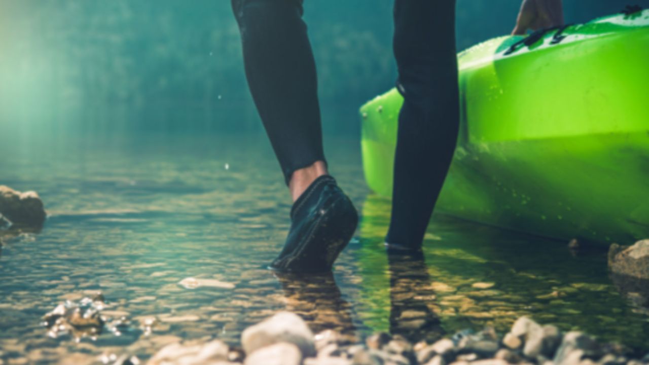 Person wearing water shoes stepping into shallow rocky water beside a kayak 