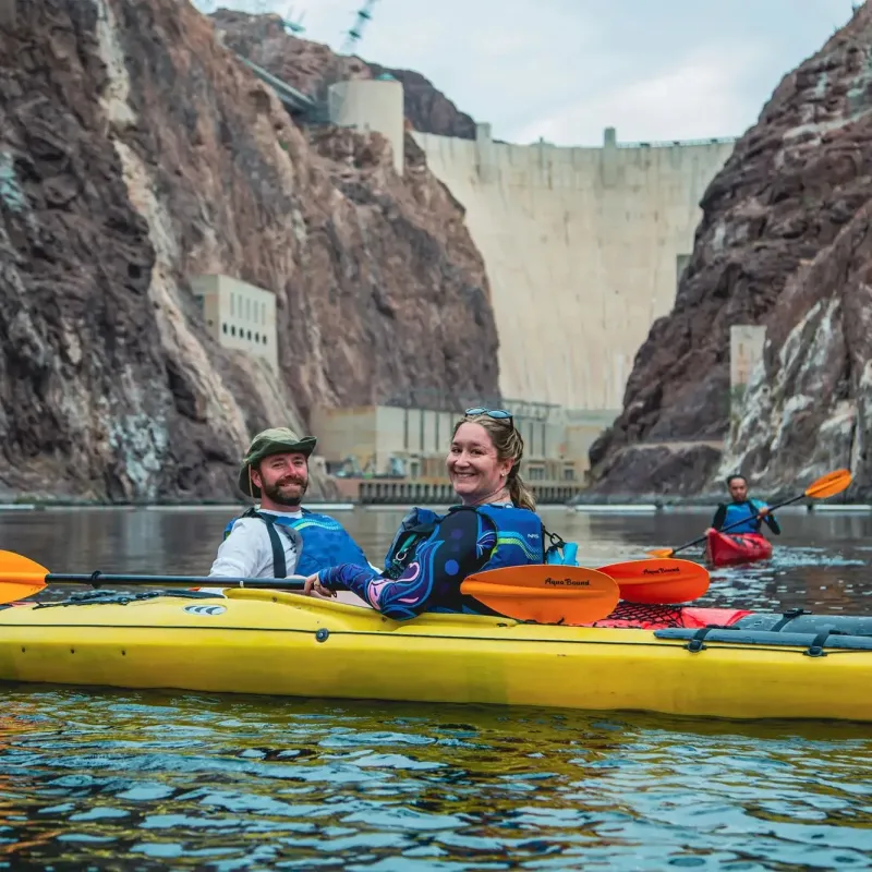 Two people kayaking on a river with a dam in the background.
