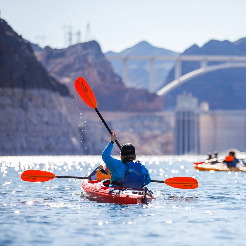 People kayaking on a river with a bridge and mountains in the background.