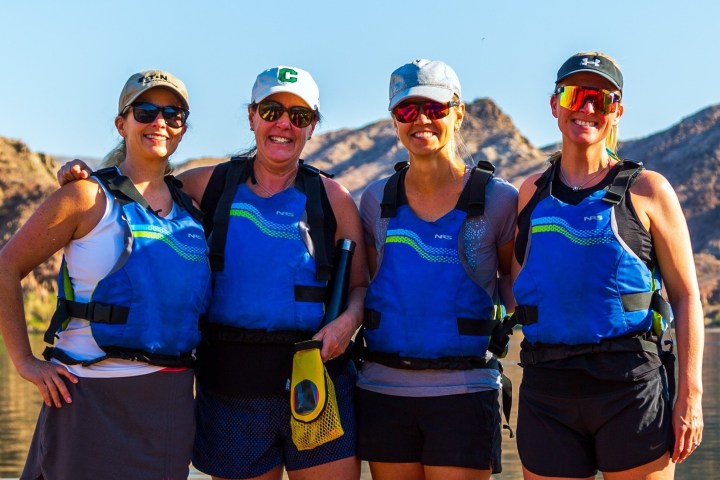 Four women in blue vests pose by a lake with hills in the background.