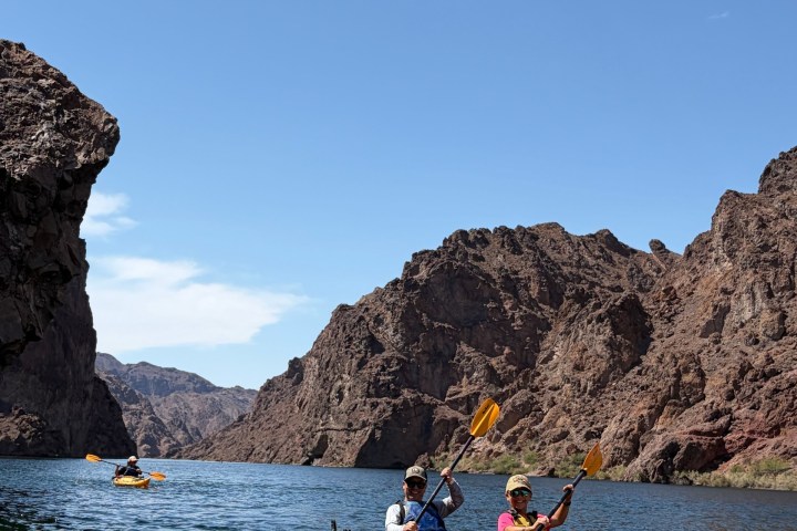 Three people kayaking on a lake surrounded by rocky cliffs under a clear blue sky.