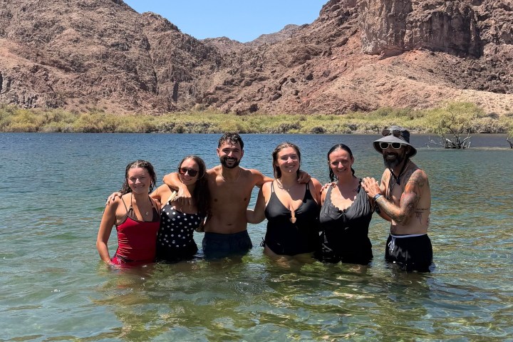 Group of people standing in clear lake with rocky hills in background.