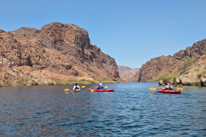 Three people kayaking on a river surrounded by rocky cliffs under a clear blue sky.