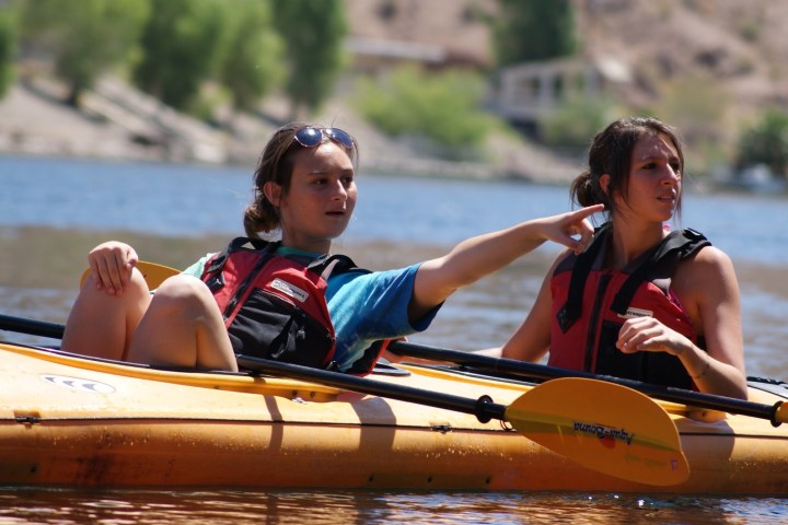 Two people in kayaks on a river, one pointing and both wearing life jackets.