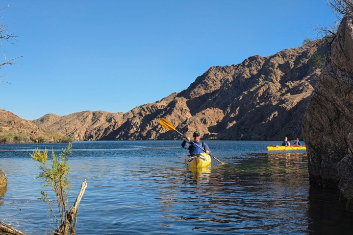 Kayakers paddle on a calm lake with rocky hills under a clear blue sky.