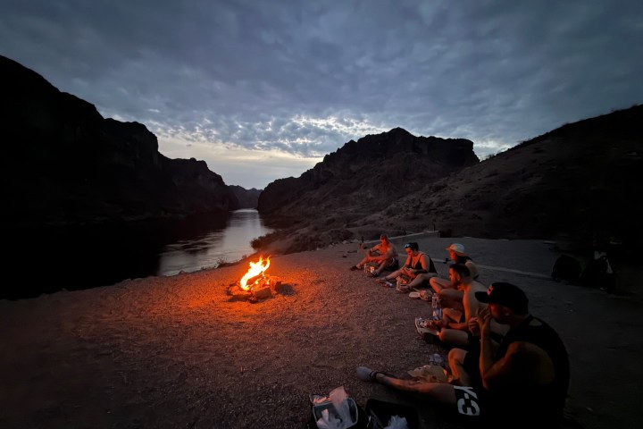 People sitting by a campfire near a river at dusk, surrounded by mountains.