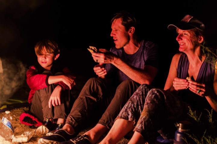Family of four sitting by a campfire at night, smiling and eating.